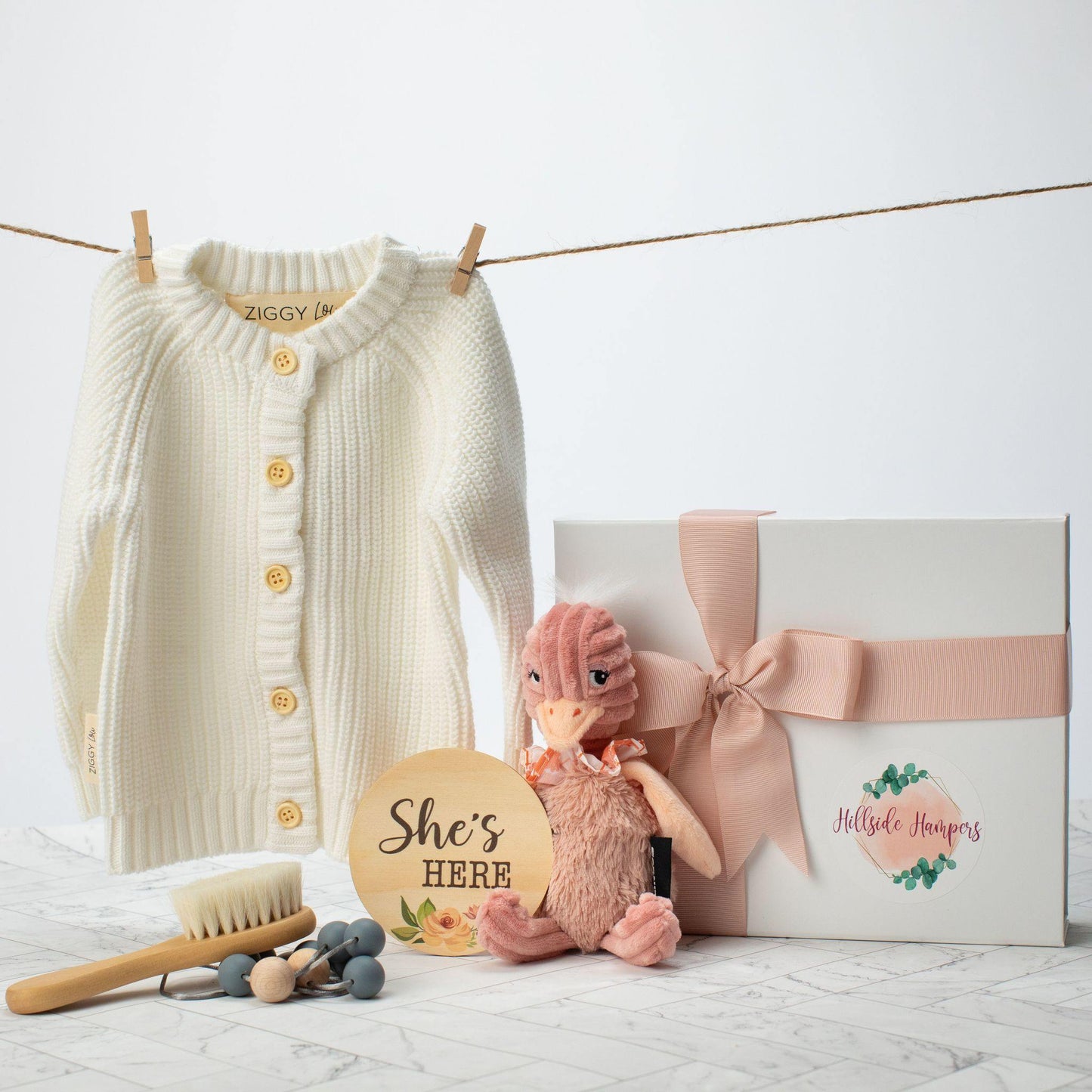 White baby cardigan next to a white gift box and soft baby toy, She's here sign, baby hair brush and teething necklace.