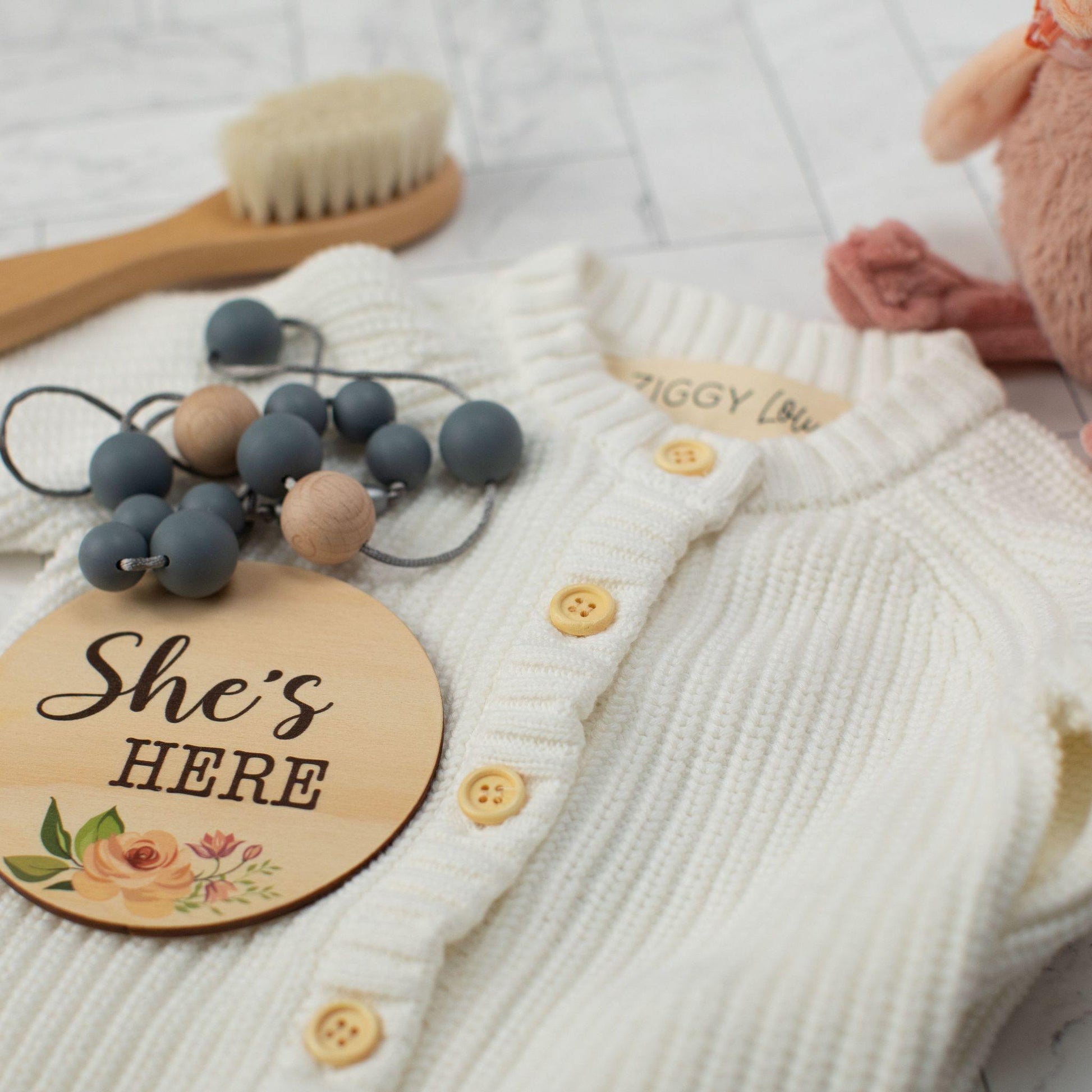 white baby cardigan with "she's here" sign and teething necklace laying on top. Baby hairbrush in the background