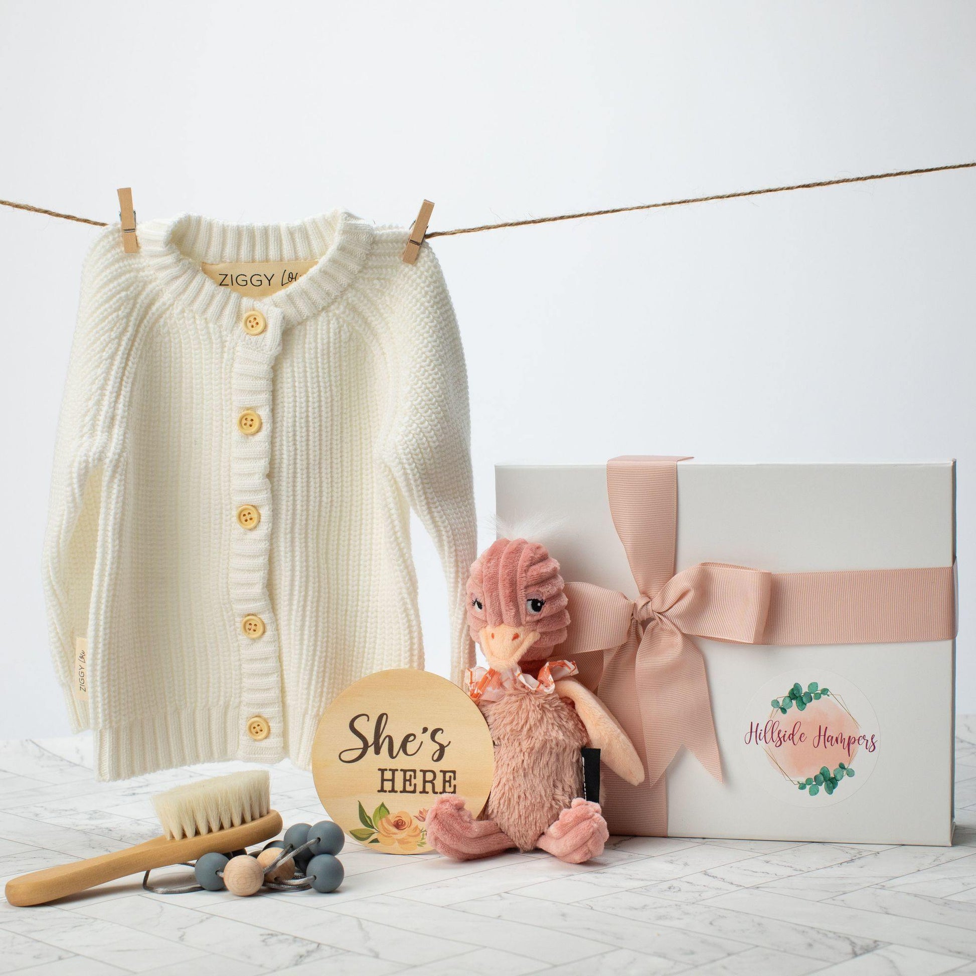 White baby cardigan next to a white gift box and soft baby toy, She's here sign, baby hair brush and teething necklace. 